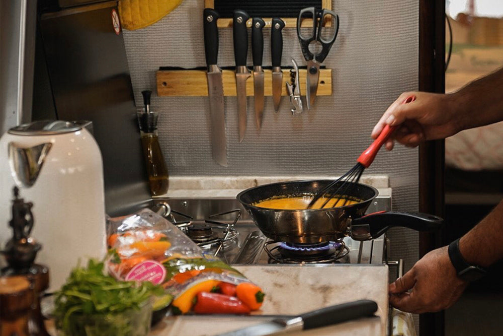 Person cooking in a small kitchen, whisking sauce in a pan on the stove, with vegetables and kitchen tools nearby.