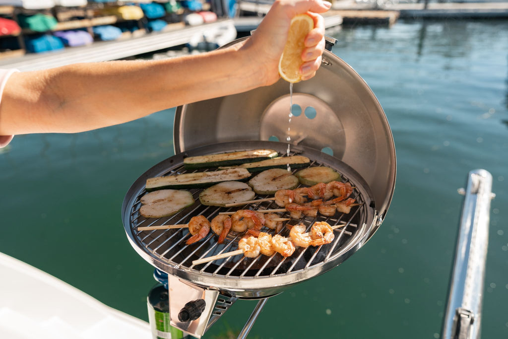 Person grilling shrimp and vegetables on a boat grill, squeezing lemon juice over them.